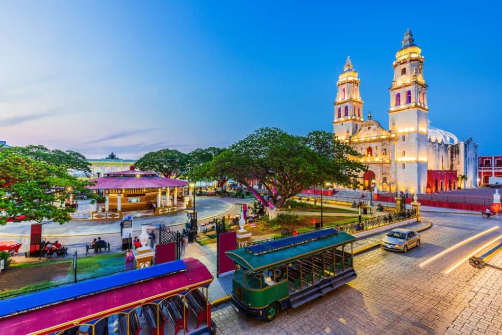 De verlichte witte kathedraal van Campeche met twee torens is te zien achter kleurrijke trolleybussen bij schemering
