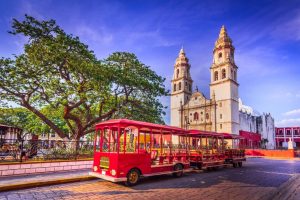 Een rode toeristentrolley staat geparkeerd voor de witte kathedraal met twee torens in het historische centrum van Campeche