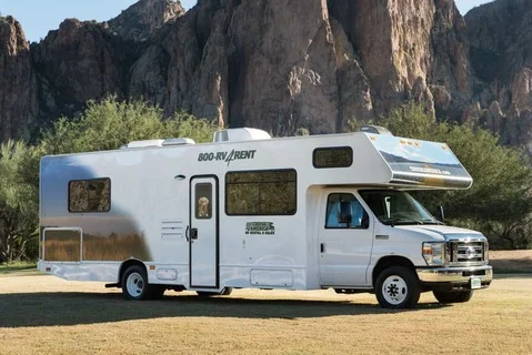 Witte camper met een opdruk van een hond op de zijkant, geparkeerd op gras met scherpe rotspieken op de achtergrond