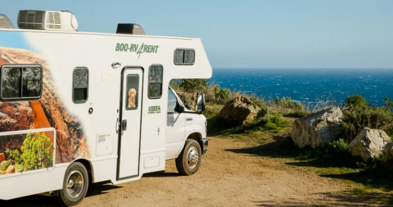Witte huurcamper geparkeerd op een onverhard pad boven een rotsachtige kust met uitzicht over de blauwe oceaan