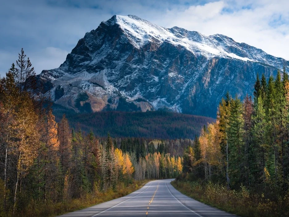 Een lege weg slingert door een herfstachtig bos met een besneeuwde bergtop op de achtergrond in Canada
