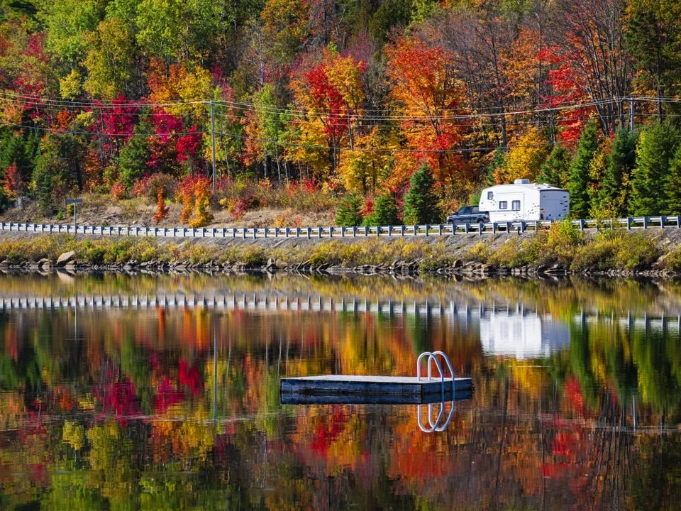 Een camper rijdt over een weg langs een meer waarin bomen met felrode en oranje herfstbladeren weerspiegelen in Canada