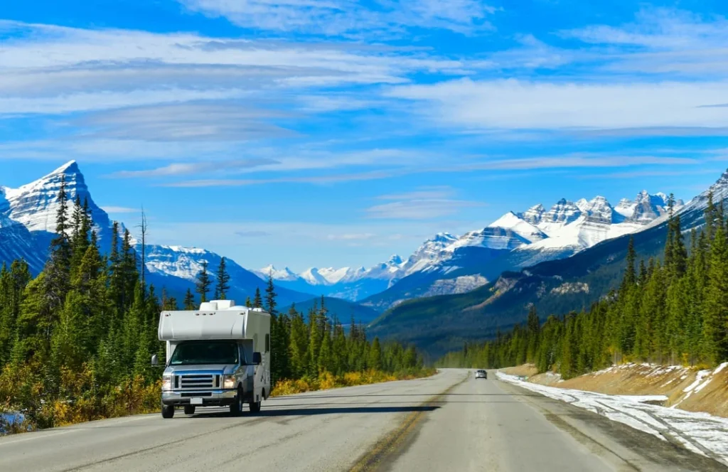 Een witte camper rijdt over een lege weg tussen dennenbossen, met besneeuwde bergtoppen op de achtergrond in Canada