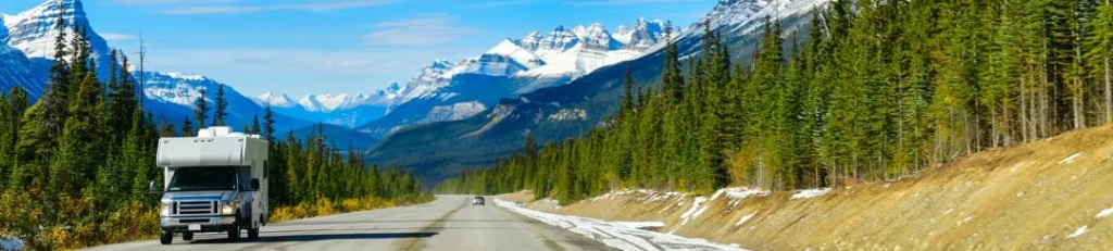 Een camper rijdt over een lege weg door de Canadese Rocky Mountains, omringd door dennenbossen en besneeuwde bergtoppen