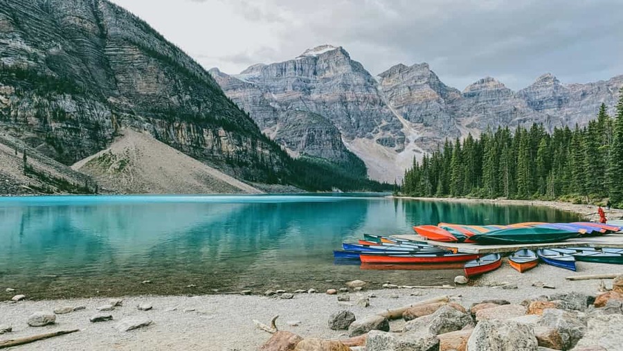 Rij gekleurde kano's op een kiezelstrand bij een turkoois meer met besneeuwde bergen op de achtergrond in Banff National Park