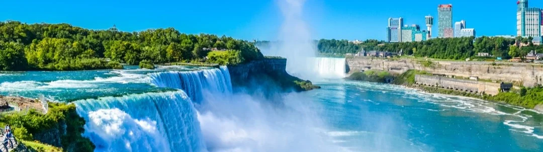 Panoramisch uitzicht op de bruisende Niagara watervallen met groen langs de oevers en de skyline van de stad op de achtergrond