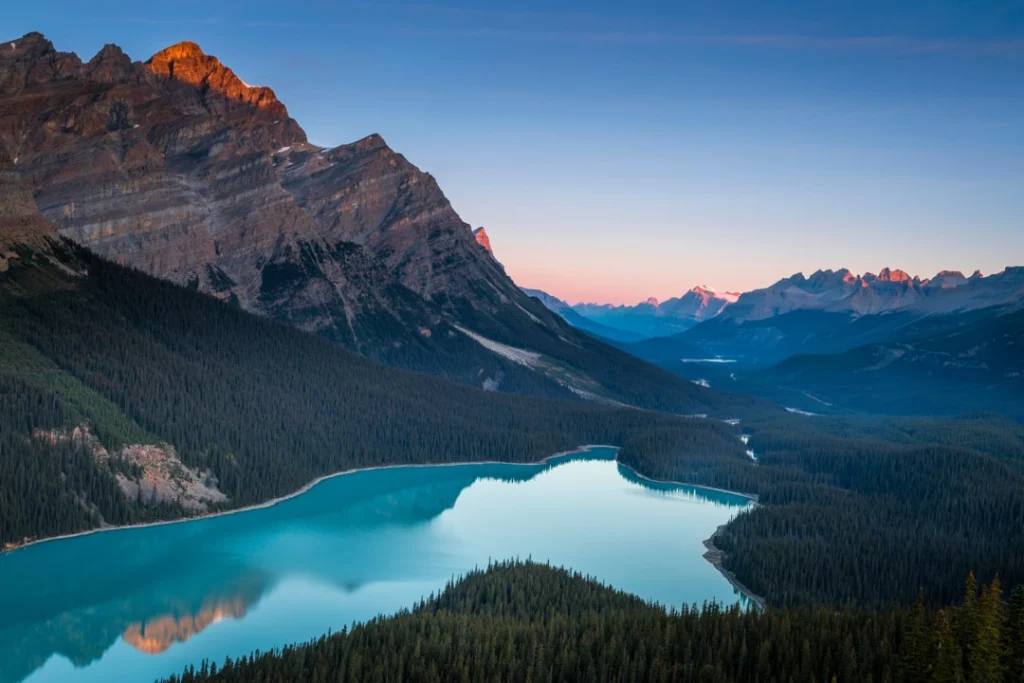 Uitzicht over het turquoise Peyto Lake in Canada, omgeven door dennenbossen en bergen die oplichten bij zonsopgang