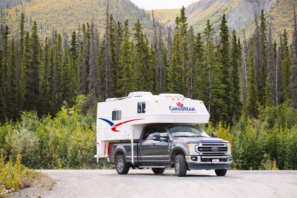 Een grijze pickup truck met camperopbouw geparkeerd op een grindweg tussen dennenbossen en bergen in West-Canada