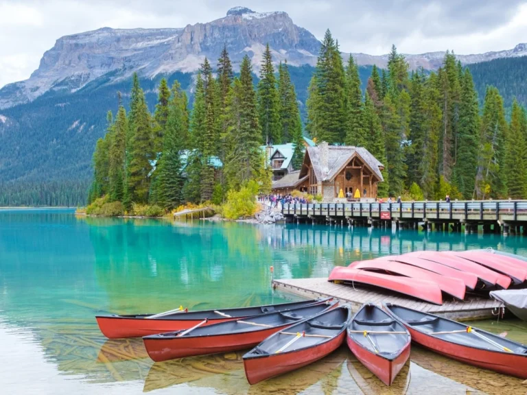 Een rij rode kano's ligt op de steiger van een turkoois bergmeer in Canada, met een houten lodge en besneeuwde bergen op de achtergrond