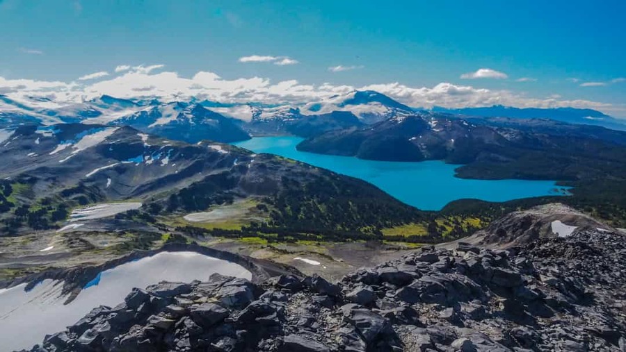 Uitzicht vanaf een rotsachtige bergtop in Whistler, Canada, over een turkooizen gletsjermeer omringd door besneeuwde bergtoppen