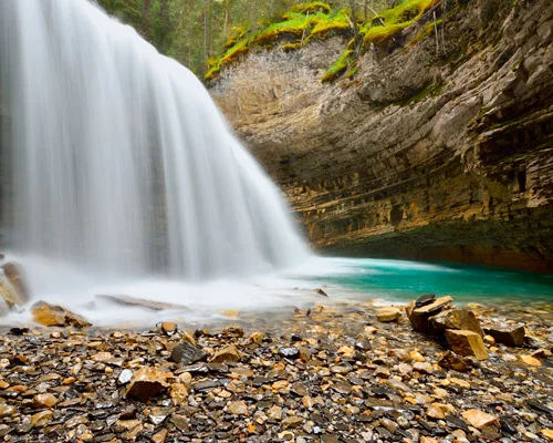 Brede waterval valt van een rotsrichel met mos naar een turquoise poel omgeven door kiezelstenen en een steile rotswand