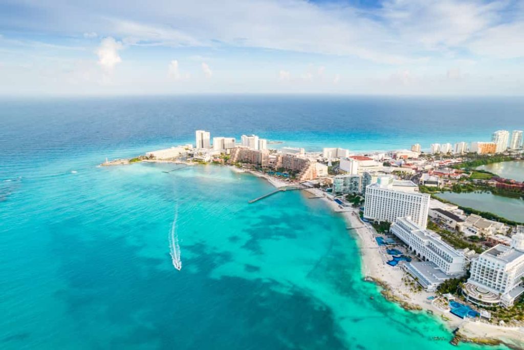 Luchtfoto van hotels langs een wit strand met een bootje dat door het heldere turkoois water van Cancún vaart