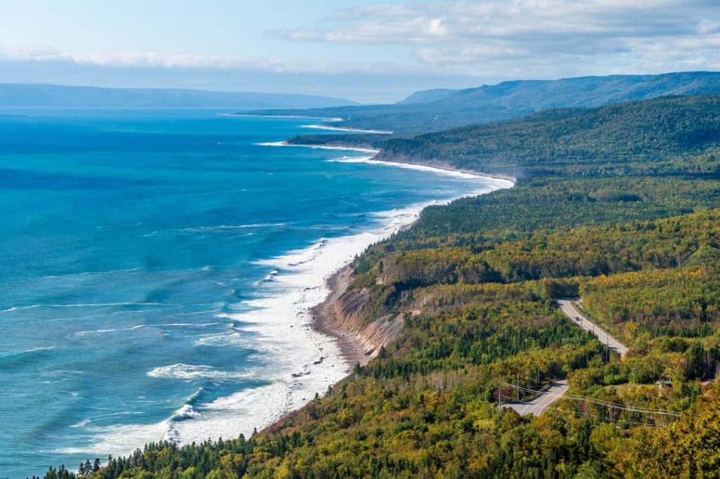 Uitgestrekte kustlijn met witte golven die breken op het strand, omringd door beboste bergen op Cape Breton