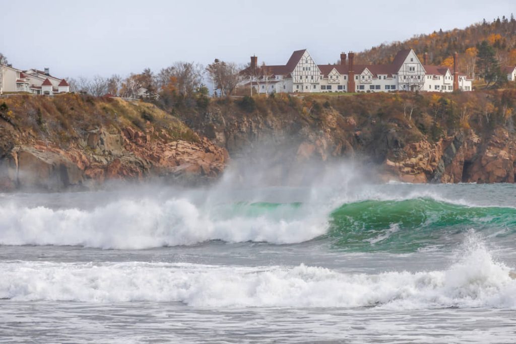 Grote schuimende golven breken op het strand onder de rotsen bij het witte hotel Keltic Lodge op Cape Breton