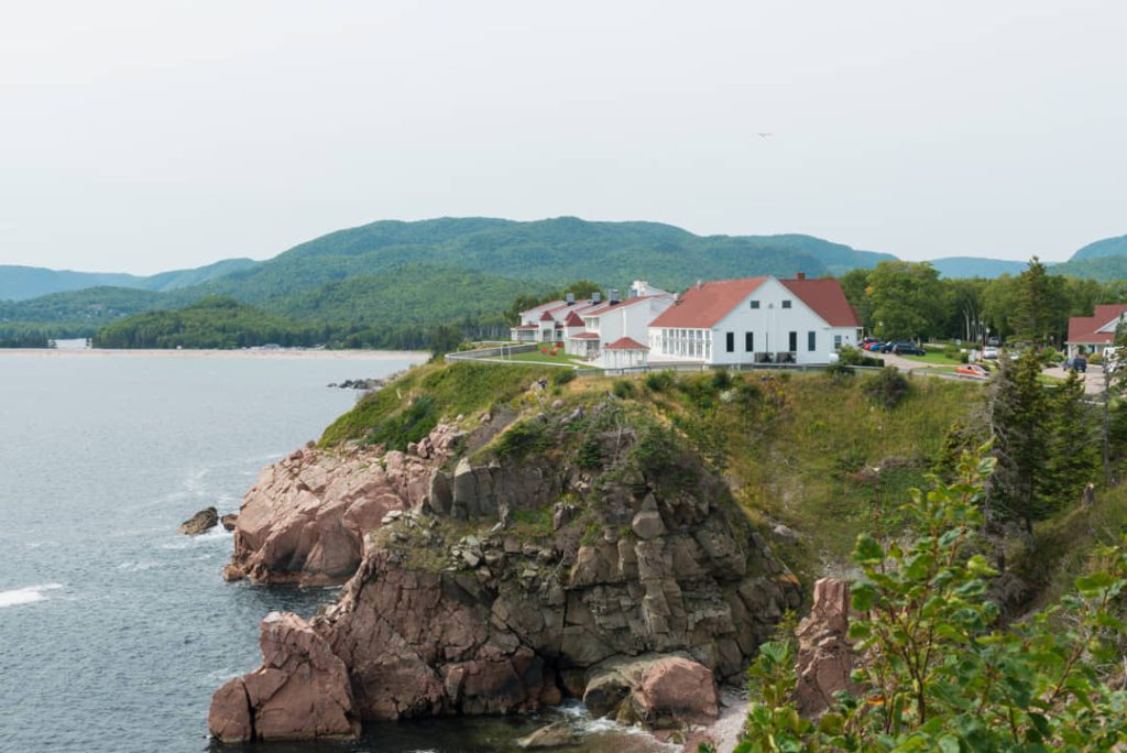 Wit hotelgebouw met rood dak op een rotsachtige landtong met beboste bergen op de achtergrond bij Cape Breton