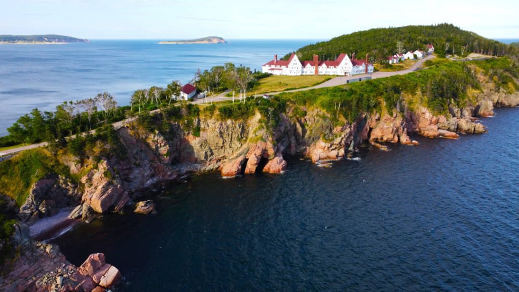 Wit hotel met rode daken gelegen op een groene landtong tussen roze rotsen en de blauwe zee bij Cape Breton