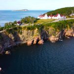 Wit hotel met rode daken gelegen op een groene landtong tussen roze rotsen en de blauwe zee bij Cape Breton