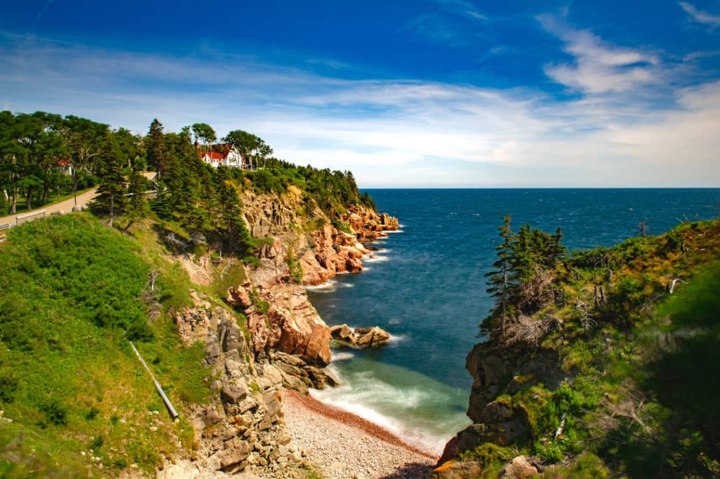 Rode rotsachtige kliffen met bos en een wit hotel op de heuvel langs de kustweg naar Keltic Lodge op Cape Breton