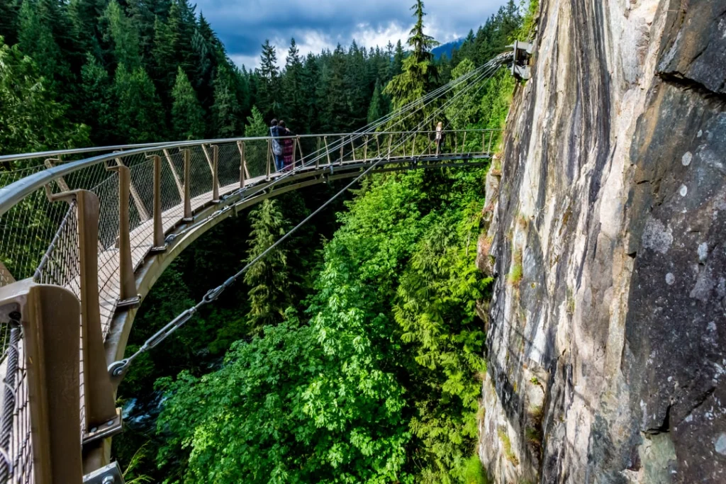 Bezoekers lopen over een smalle hangbrug die tegen een rotswand is bevestigd, hoog boven een groen ravijn bij de Capilano Cliff Walk