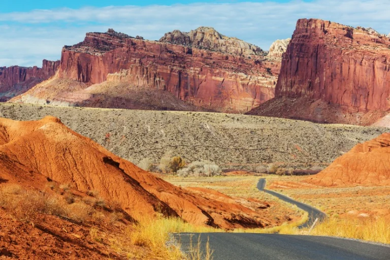 Een smalle asfaltweg slingert tussen droog struikgewas en hoge rode zandstenen rotswanden in Capitol Reef National Park, Utah