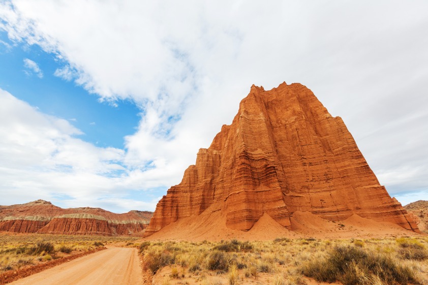Een grote gelaagde rode rotstoren torent boven een onverharde weg met struikgewas in Capitol Reef National Park