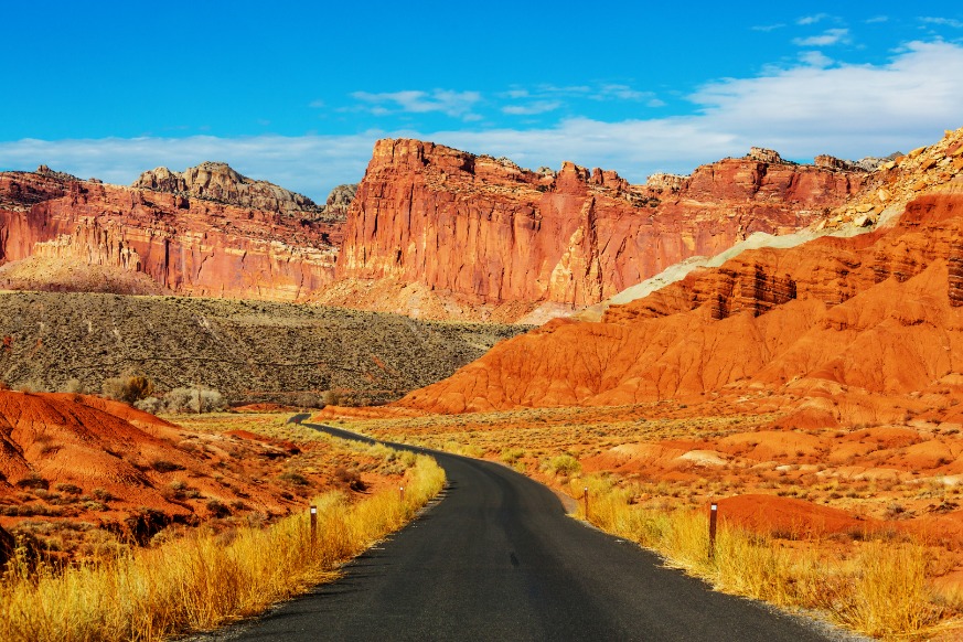 Een smalle weg loopt tussen droog struikgewas naar hoge, gelaagde rode rotswanden onder een blauwe hemel in Capitol Reef National Park