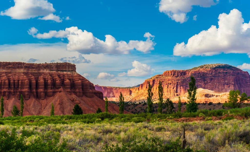 Gelaagde rode rotsformaties met groene bomen op de voorgrond onder een blauwe lucht met wolken in Capitol Reef National Park