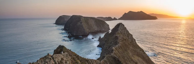 Smalle rotsachtige bergkam steekt uit in de oceaan onder een warme avondzon met eilanden op de achtergrond in het Channel Islands National Park