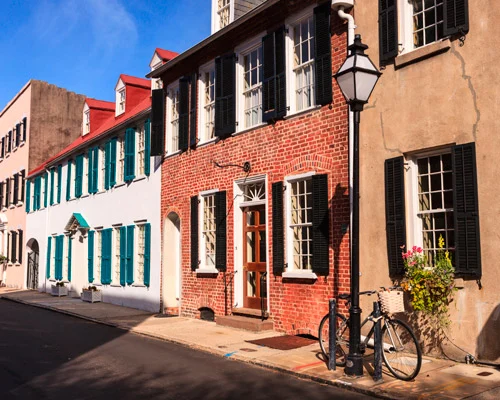 Rij historische bakstenen en gepleisterde huizen met zwarte luiken langs een straat in Charleston met een fiets tegen een lantaarnpaal