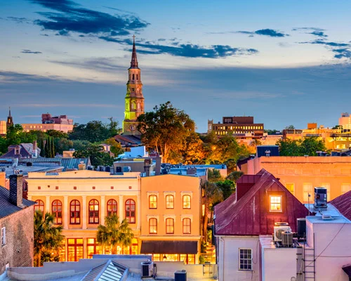 Daken en gevels van historische gebouwen met een hoge witte kerktoren op de achtergrond in Charleston in South Carolina bij het invallen van de avond