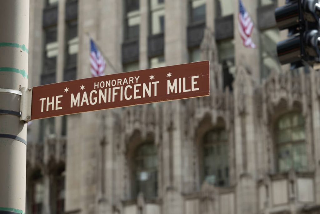 Bruin straatnaambord met de tekst The Magnificent Mile aan een lantaarnpaal met op de achtergrond de gotische gevel van een historisch gebouw en Amerikaanse vlaggen