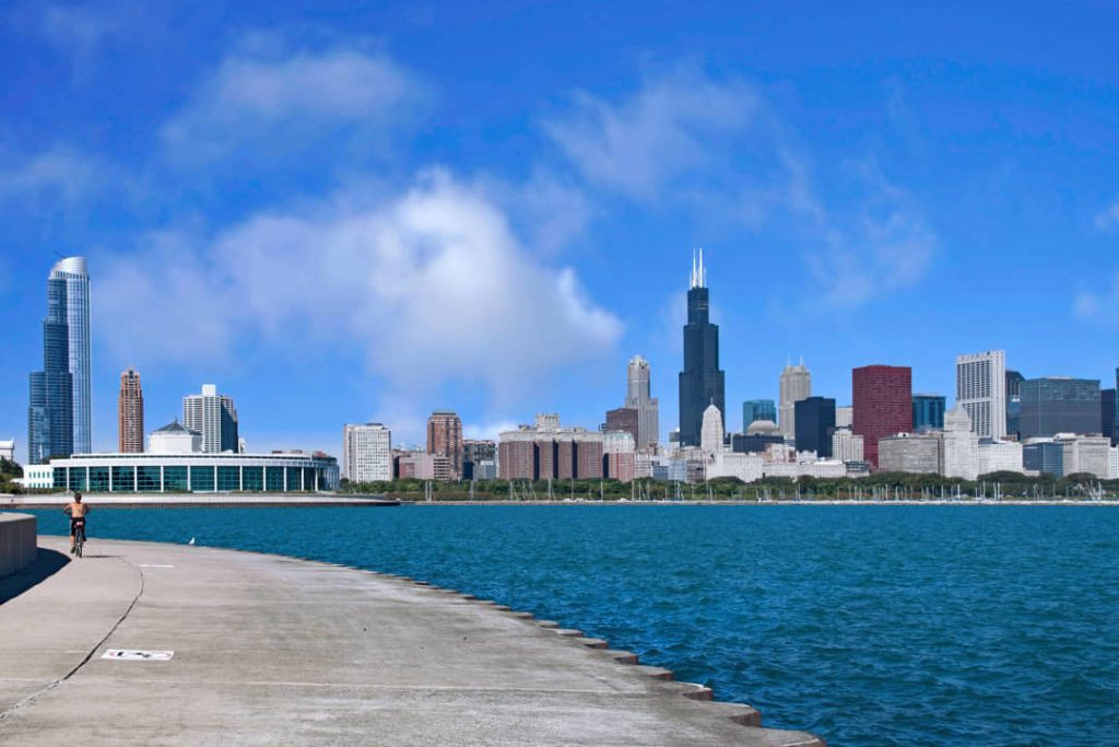 Fietser rijdt over een betonnen pad langs het meer terwijl in de verte de wolkenkrabbers van Chicago met de Willis Tower zichtbaar zijn onder een bewolkte lucht