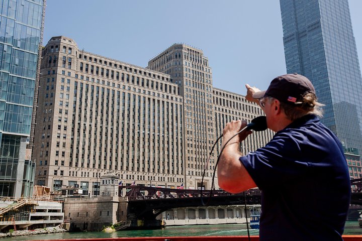 Man met pet en microfoon geeft op een rondvaartboot uitleg over een groot gebouw aan de oever van de Chicago River terwijl op de achtergrond een wolkenkrabber oprijst