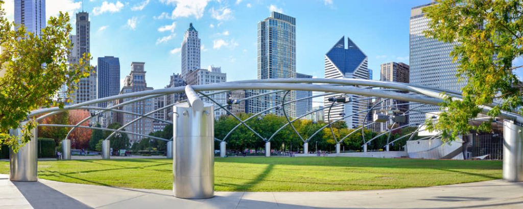 Metalen boogconstructie van het openluchttheater in Millennium Park met een groen grasveld ervoor en de skyline van Chicago op de achtergrond