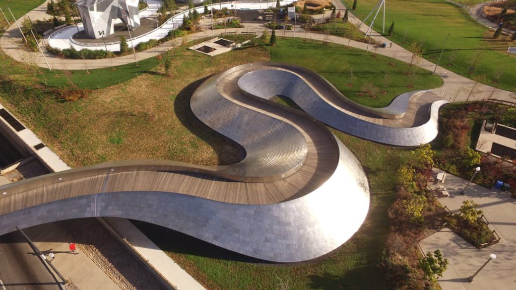 Luchtfoto van bovenaf van de kronkelende metalen voetgangersbrug in Millennium Park met een parkeergarage en groene struiken eromheen