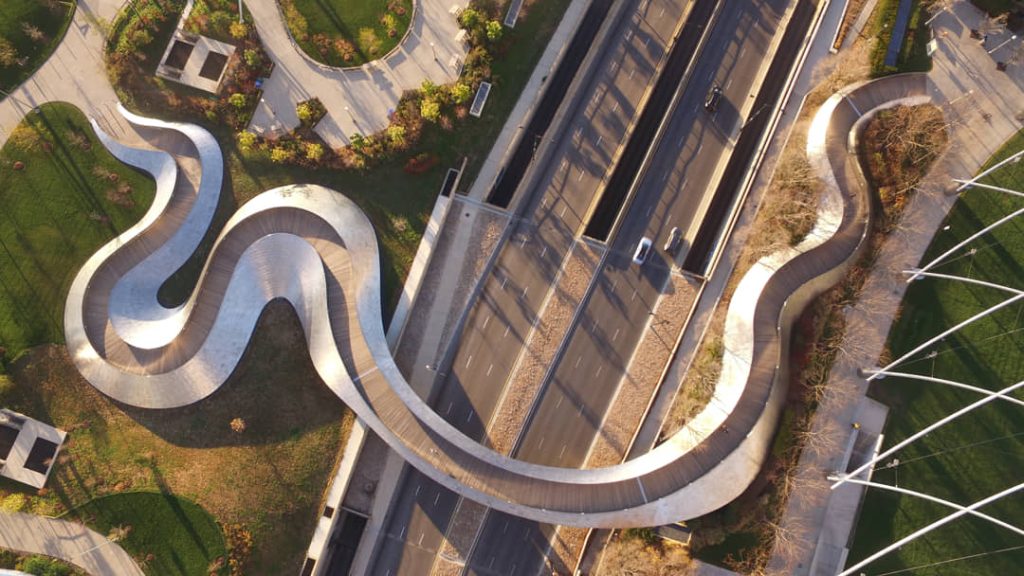 Luchtfoto recht van boven van een glanzende metalen voetgangersbrug met een slingerende vorm boven een snelweg en groen park in Chicago