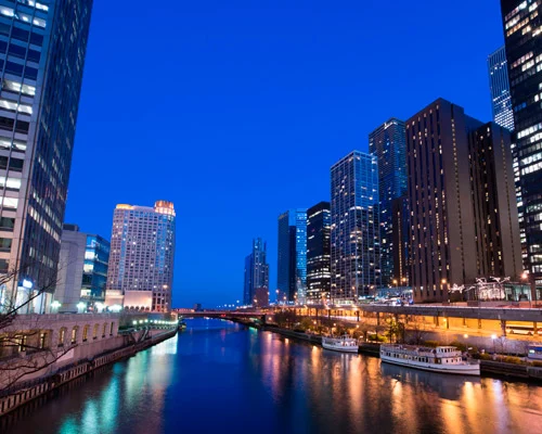 Afgemeerde boten langs de Chicago River met hoge verlichte wolkenkrabbers aan beide oevers onder een blauwe avondlucht