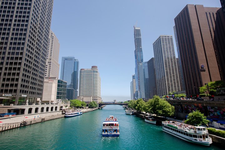 Meerdere rondvaartboten varen op het turquoise water van de Chicago River met aan weerszijden hoge kantoortorens en een heldere blauwe lucht