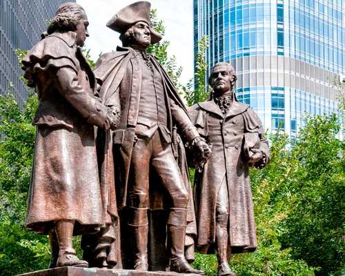Close-up van een bronzen standbeeldengroep met drie mannen in achttiende-eeuwse kleding voor wolkenkrabbers en groen gebladerte in Chicago