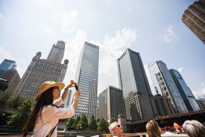 Vrouw met strooien hoed maakt op haar telefoon een foto van hoge wolkenkrabbers langs de Chicago River terwijl ze op een rondvaartboot staat