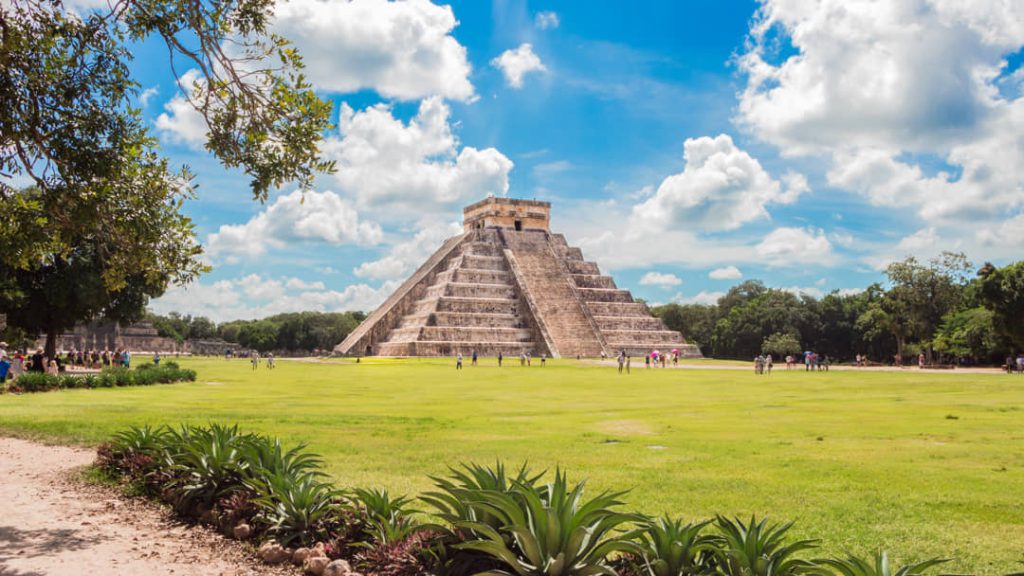 De piramide van Kukulcán is te zien tussen bomen met agaveplanten op de voorgrond en een wolkenhemel erboven