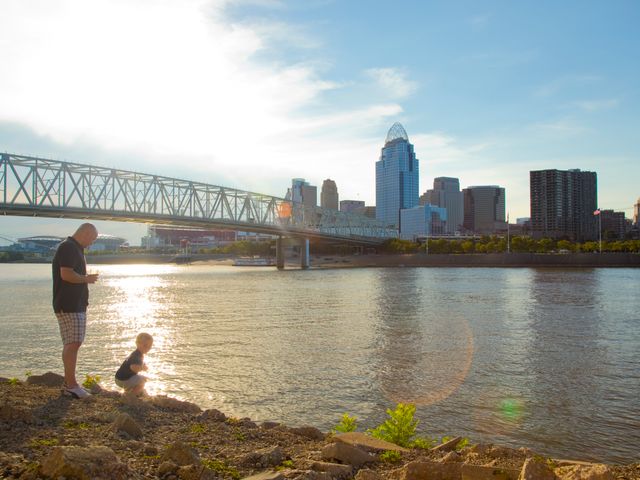 Vader en kind kijken naar de rivier in Cincinnati terwijl de skyline en de historische Roebling Suspension Bridge oplichten