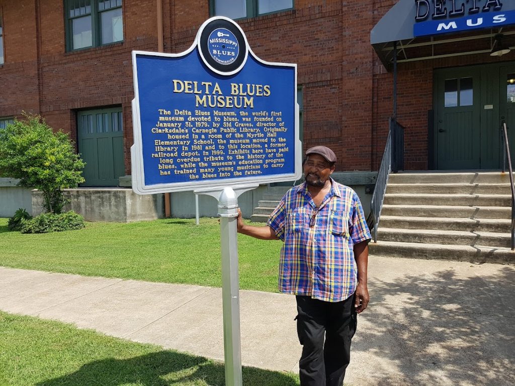 Man in geruit overhemd staat naast een blauw informatiebord over het Delta Blues Museum voor een bakstenen gebouw in Clarksdale