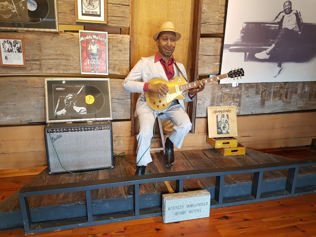 Levensgrote pop van bluesmuzikant Muddy Waters in wit pak speelt gitaar op een klein podium tussen ingelijste platen en foto's in het Delta Blues Museum