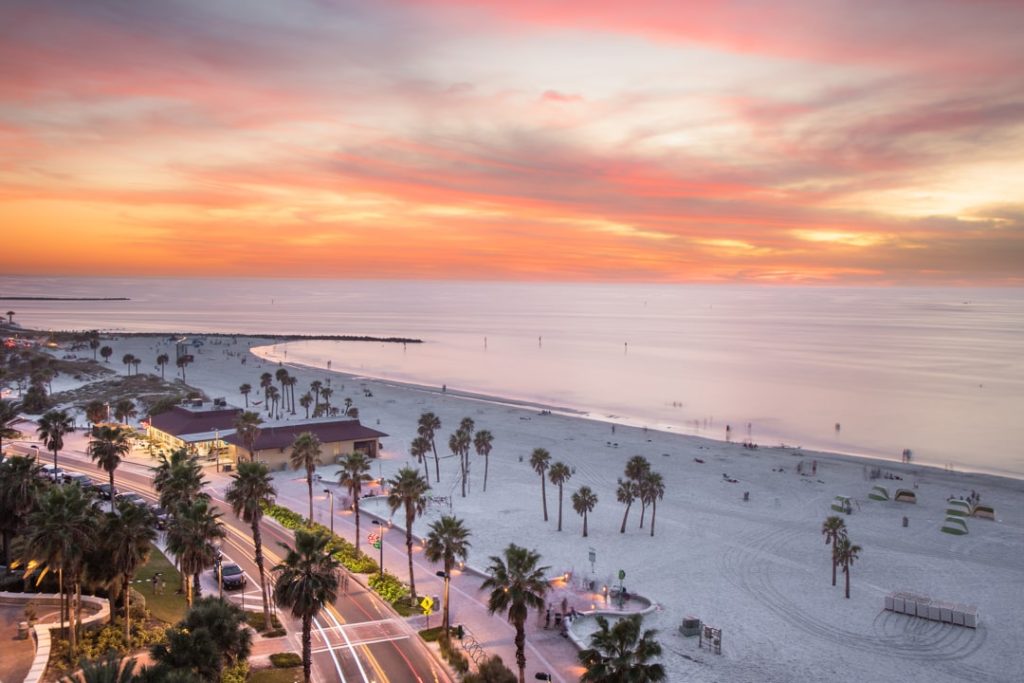 Luchtfoto van het strand van Clearwater Beach bij zonsondergang met palmbomen, een pier en een weg met lichtsporen van auto's