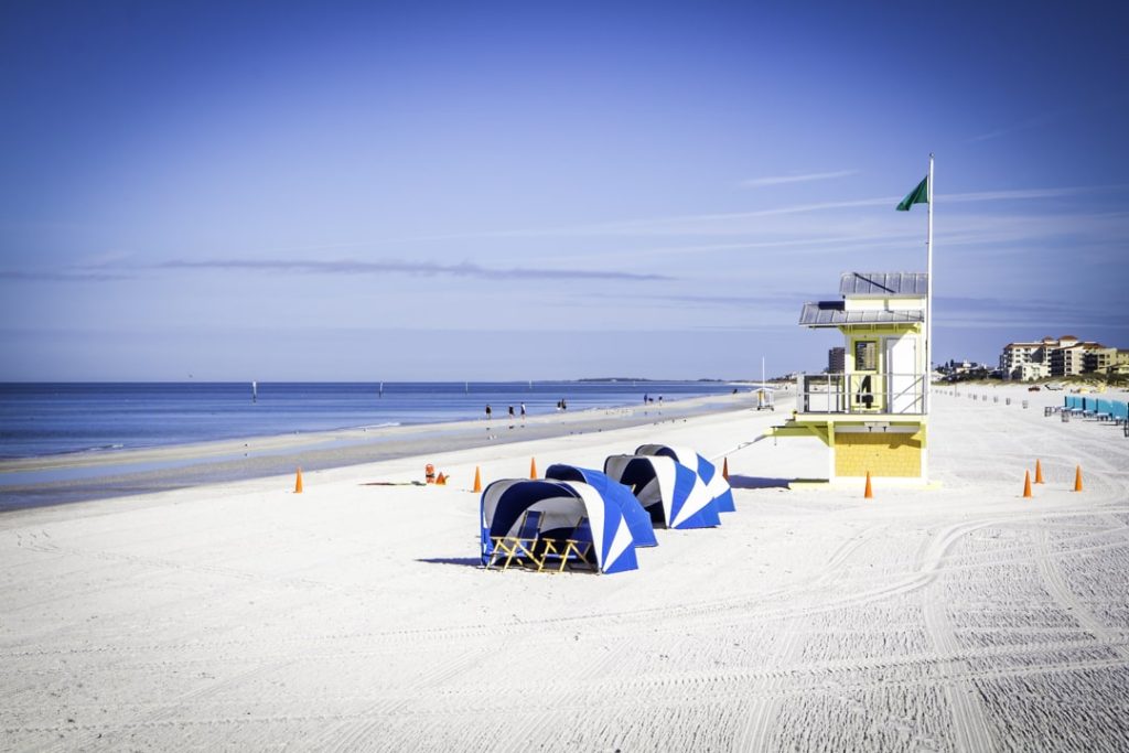 Wit zandstrand van Clearwater Beach met een gele reddingspost, blauw witte strandtenten en een vlaggenmast onder een strakblauwe hemel