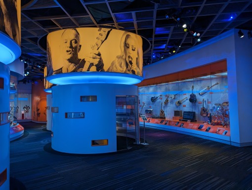 Rij gitaren tentoongesteld tegen een verlichte wand met een groot rond scherm met historische muzikanten in het GRAMMY Museum Mississippi