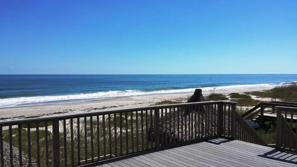 Een houten terras met trap leidt naar een breed zandstrand met golvende zee bij Cocoa Beach onder een strakblauwe hemel