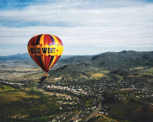 Kleurrijke heteluchtballon met de tekst Wild West vliegt boven een vallei met een klein stadje en groene bergen in Colorado