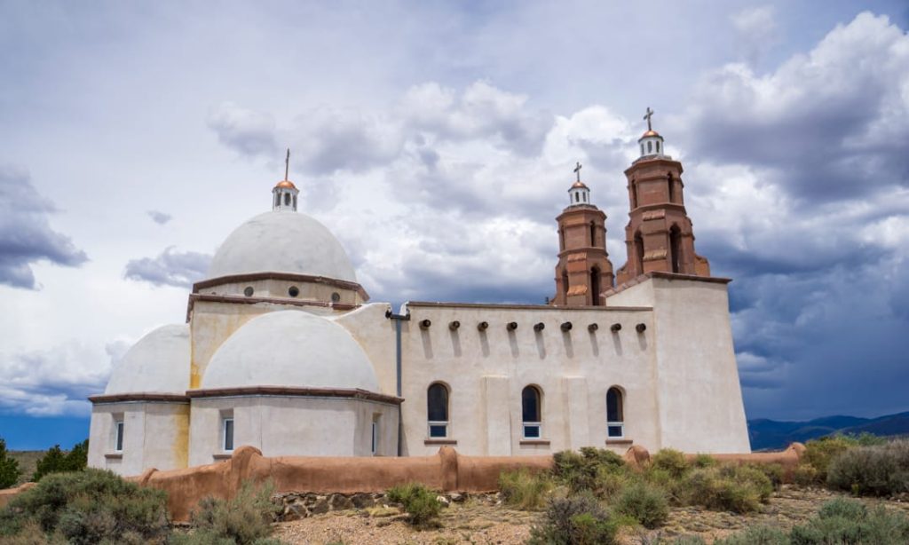 Witte adobe kerk met koepeldak en twee bakstenen torens met kruizen tegen een bewolkte lucht
